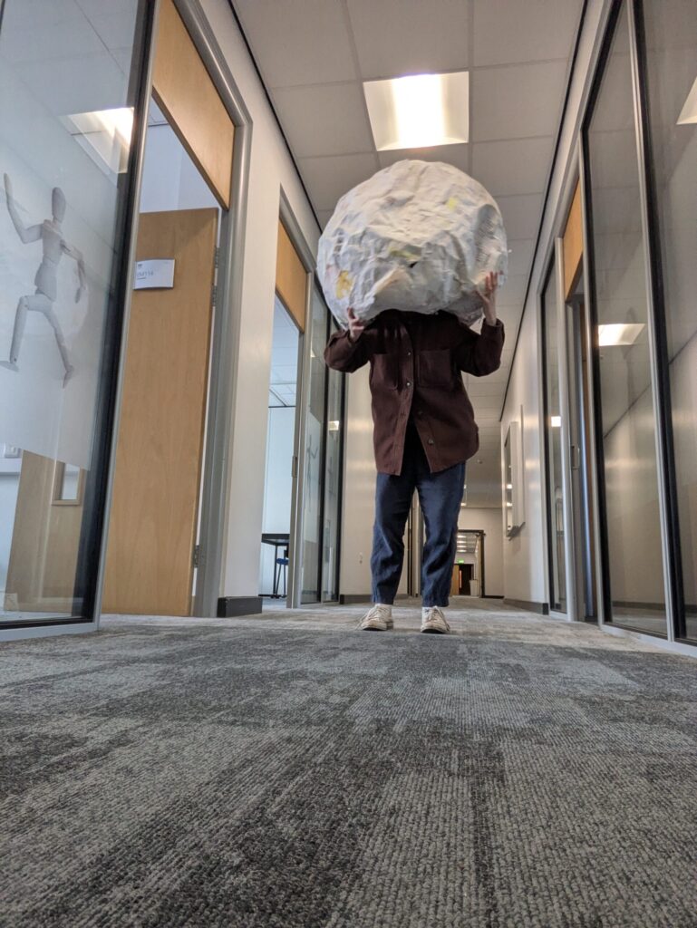 a figure in the middle ground faces the camera. They are standing in an institutional corridor. They wear a large paper sphere over their head. 